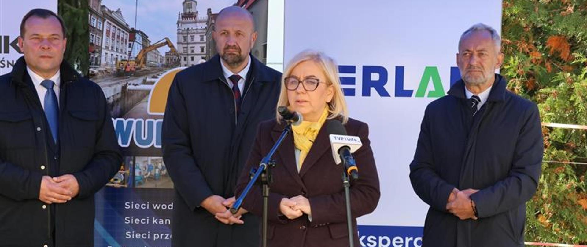 Cztery osoby stoją przed mikrofonami podczas wydarzenia plenerowego. W tle widoczne są banery informacyjne związane z inwestycjami wodno-kanalizacyjnymi oraz działalnością branżową.