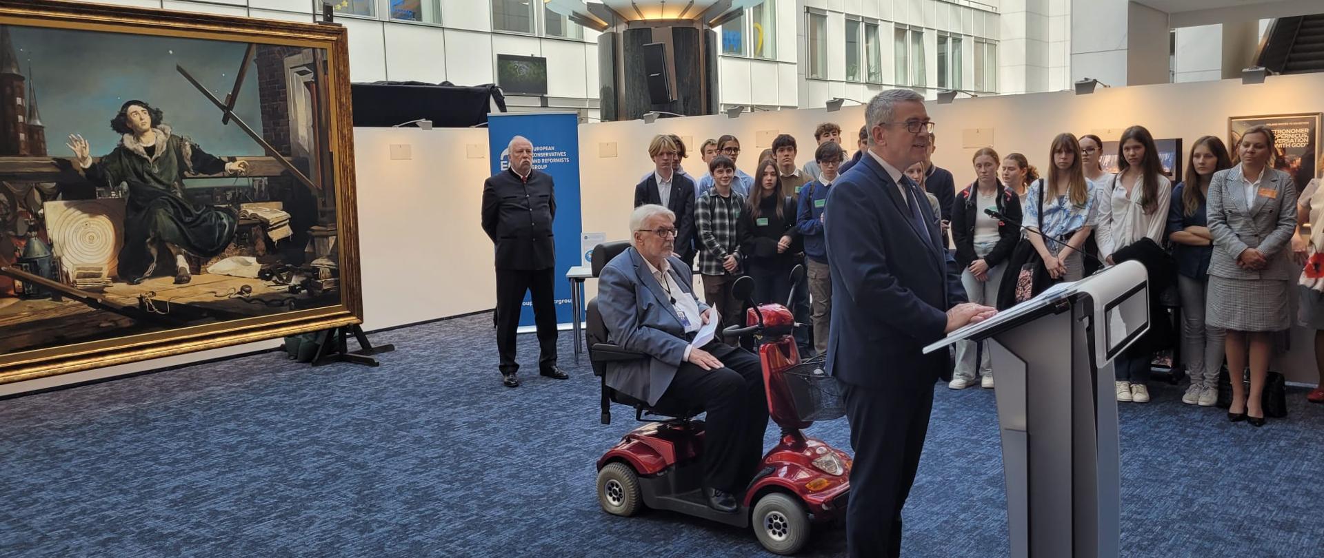 Widok na podwórko pomiędzy białymi budynkami, pod ścianą stoi obraz przedstawiający Kopernika, przed nim stoi wiceminister Murdzek i mówi do mikrofonu na stojaku, za nim mężczyzna na czterokołowym wózku inwalidzkim, w tle grupa ludzi.