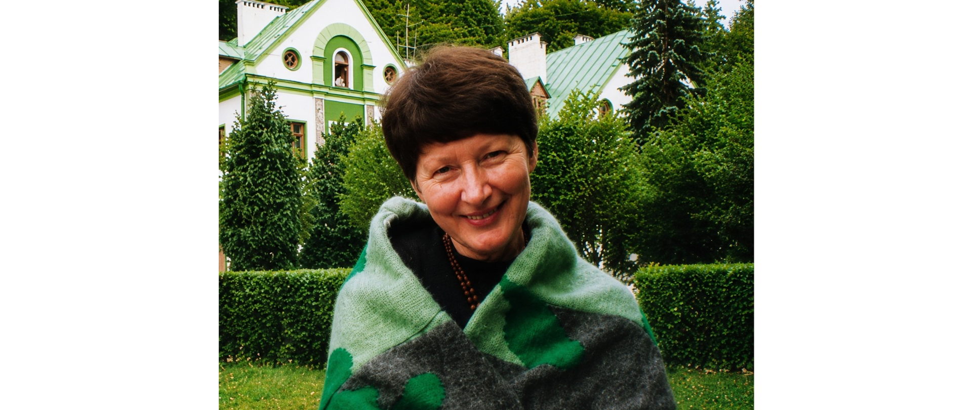 Grafika przedstawia uśmiechniętą kobietę na tle drzew oraz biało-zielonych domów.