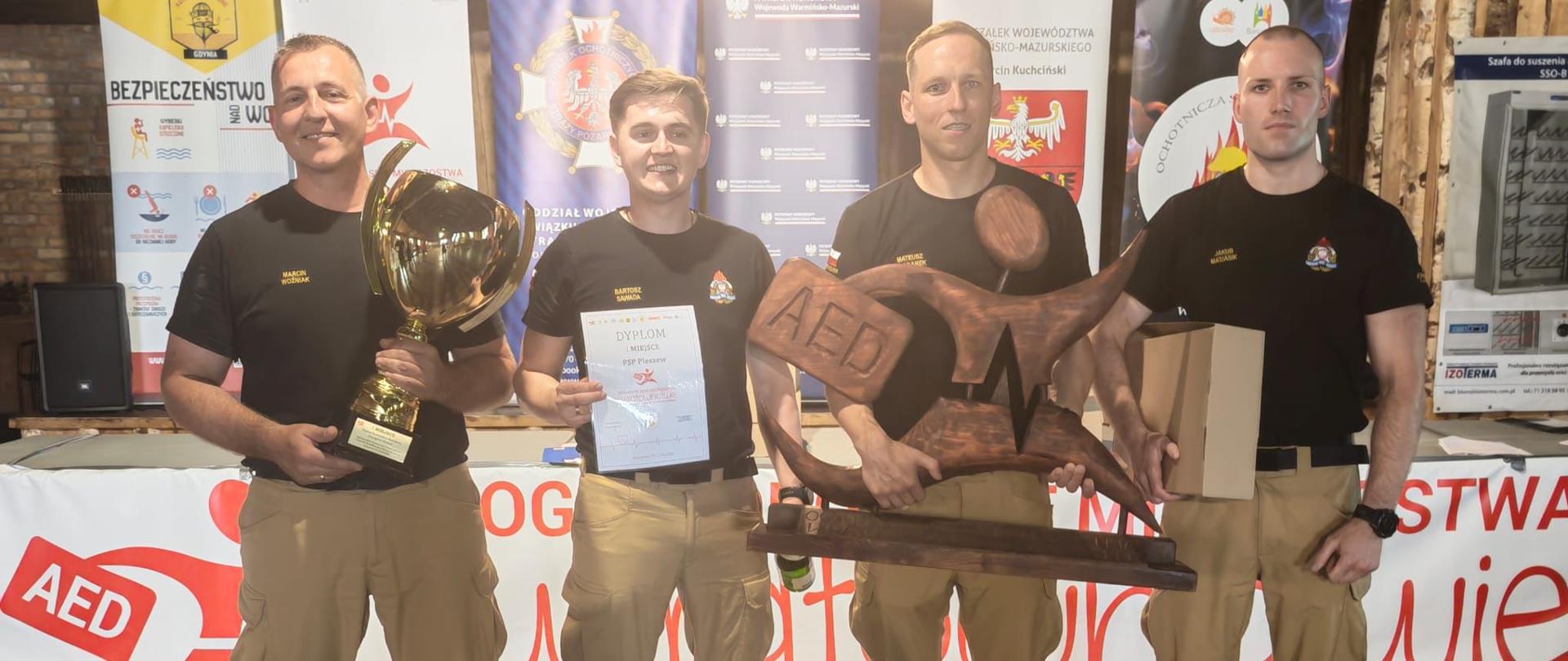 czterech strażaków z pucharem, dyplomem oraz rzeźbą stojący frontem do obiektywu