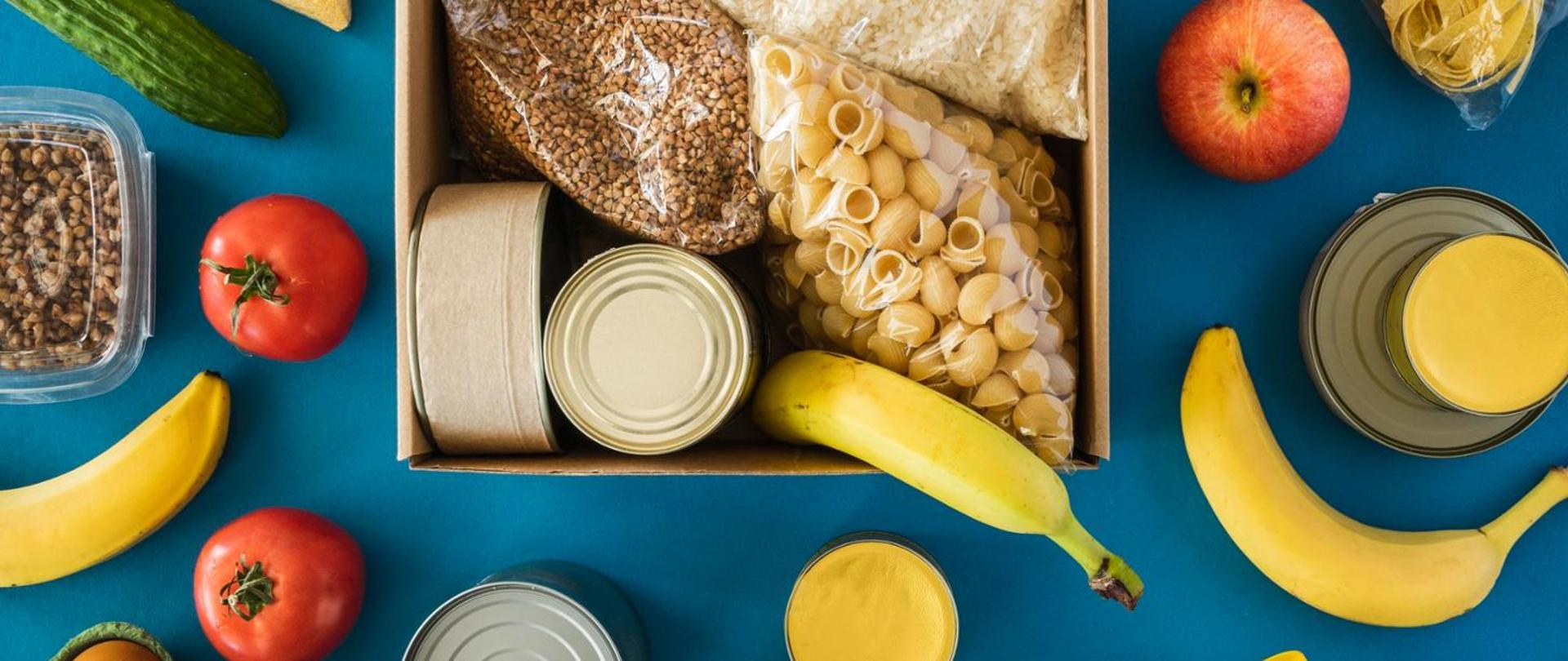W centralnej części obrazu na niebieskim tle znajduje się otwarte pudełko z umieszczonymi w środku produktami spożywczymi (paczka makaronu, opakowanie kaszy i ryżu, metalowe puszki i banan). Wokół pudełka leżą pojedyncze owoce (czerwone jabłko i banany), warzywa (ogórek i pomidory), a także opakowania z makaronem i kaszą i konserwy.