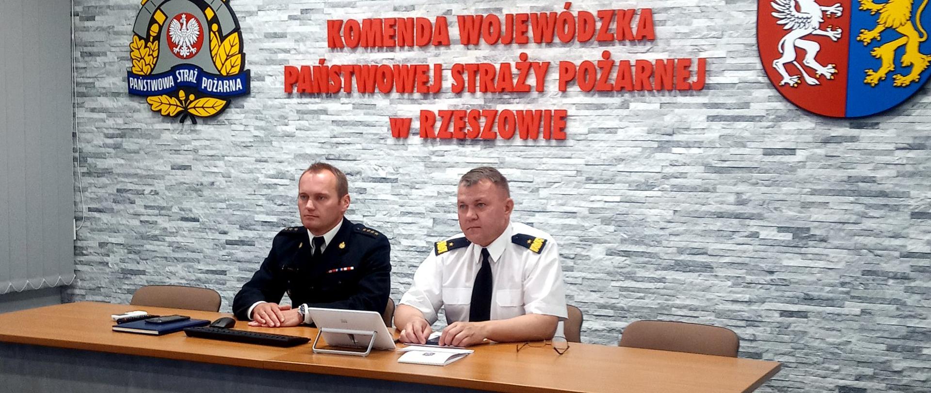 Zdjęcie zrobione w sali konferencyjnej. Na zdjęciu dwóch oficerów Państwowej Straży Pożarnej siedzi za stołem konferencyjnym. Strażak po prawej stronie w białej koszuli wyjściowej z dystynkcjami nadbrygadiera, po lewej stronie strażak w mundurze wyjściowy w stopniu starszego kapitana. Za strażakami na ścianie napis Komenda Wojewódzka Państwowej Straży Pożarnej w Rzeszowie, po lewej stronie logo PSP, po prawej herb województwa podkarpackiego.