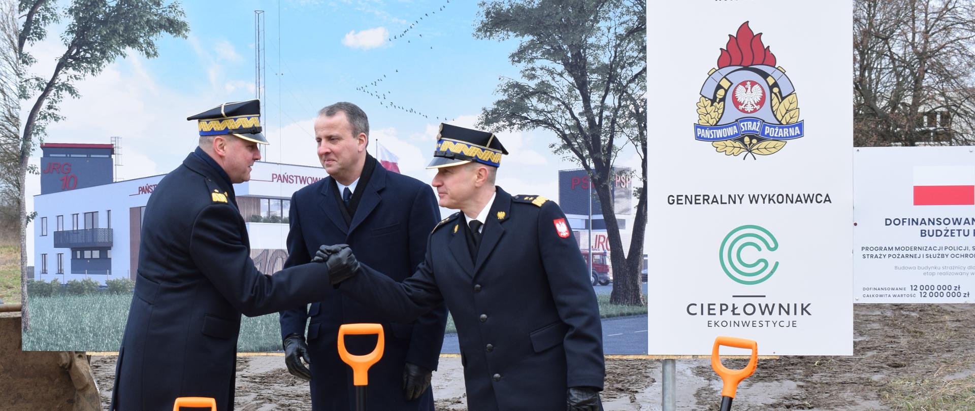 dwóch strażaków w płaszczach i rogatywkach ściskają dłoń, pomiędzy nimi stoi uśmiechnięty mężczyzna, w tle duży obraz budynku i koparka, plac budowy