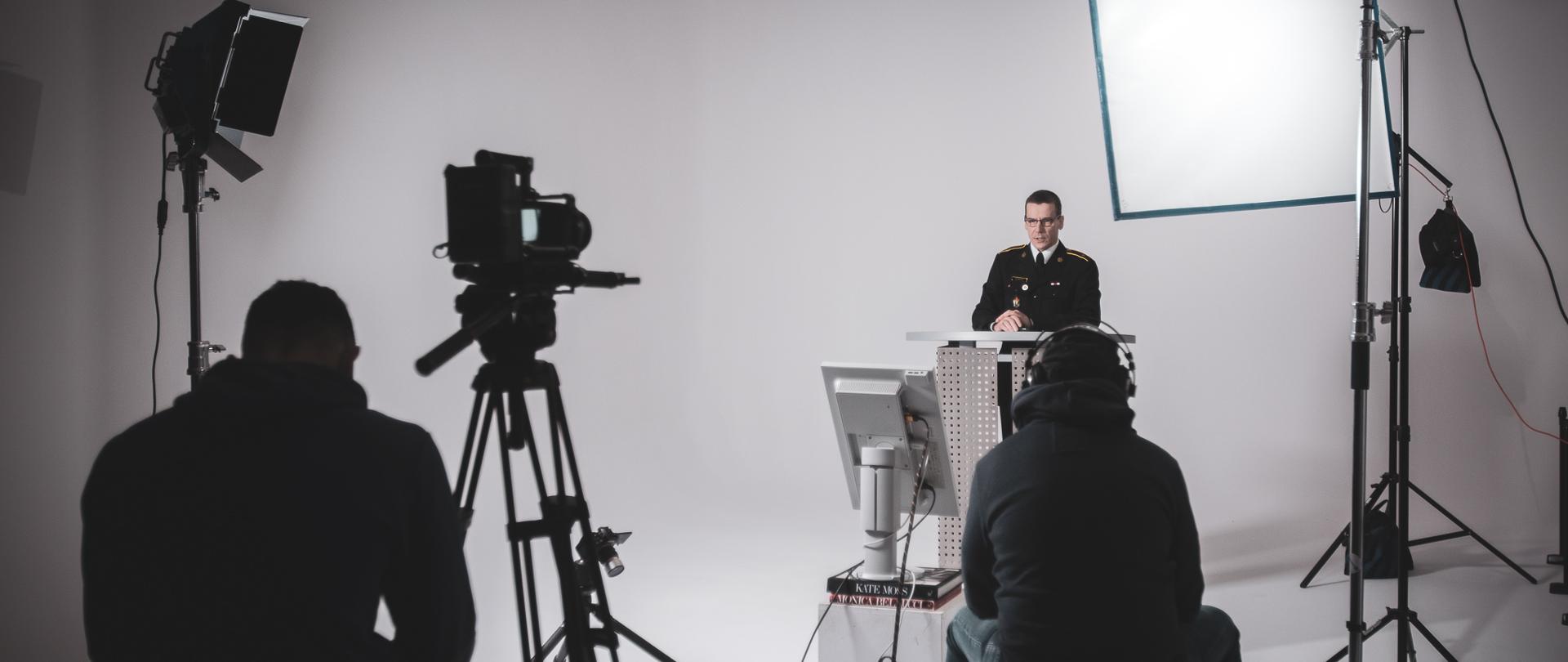 W studio filmowym z lewej strony przy mównicy stoi mężczyzna. Na przeciwko jeden mężczyzna z prawej strony obsługuje oświetlenie drugi mężczyzna z lewej strony obsługuje kamerę oraz robi zdjęcia. 