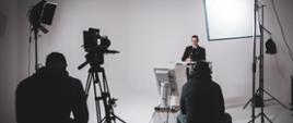 W studio filmowym z lewej strony przy mównicy stoi mężczyzna. Na przeciwko jeden mężczyzna z prawej strony obsługuje oświetlenie drugi mężczyzna z lewej strony obsługuje kamerę oraz robi zdjęcia. 