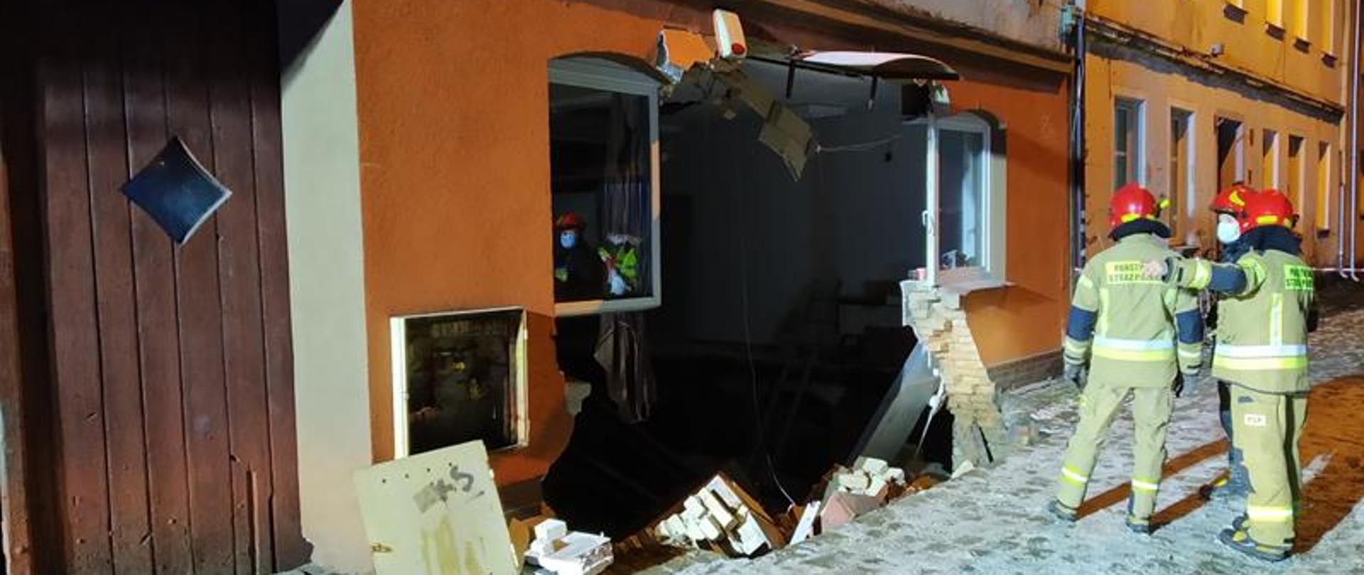 Zdjęcie przedstawia widok zawalonej ściany frontowej pierwszej kondygnacji nadziemnej budynku mieszkalnego, widoczna zabudowa szeregowa, ujęcie z lewej strony ściany. Po prawej stronie zdjęcia kierujący działaniem ratowniczym wydaje polecenia dwóm ratownikom PSP.