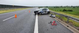 Droga ekspresowa S5. Częściowo na pasie awaryjnym stoi samochód uczestniczący w kolizji, koloru srebrnego. Za nim bariera energochłonna i teren zielony. Samochód ma uszkodzony przód oraz bok od strony kierowcy, obok leży oderwany zderzak. Na jezdni rozstawione są pachołki i trójkąt ostrzegawczy.