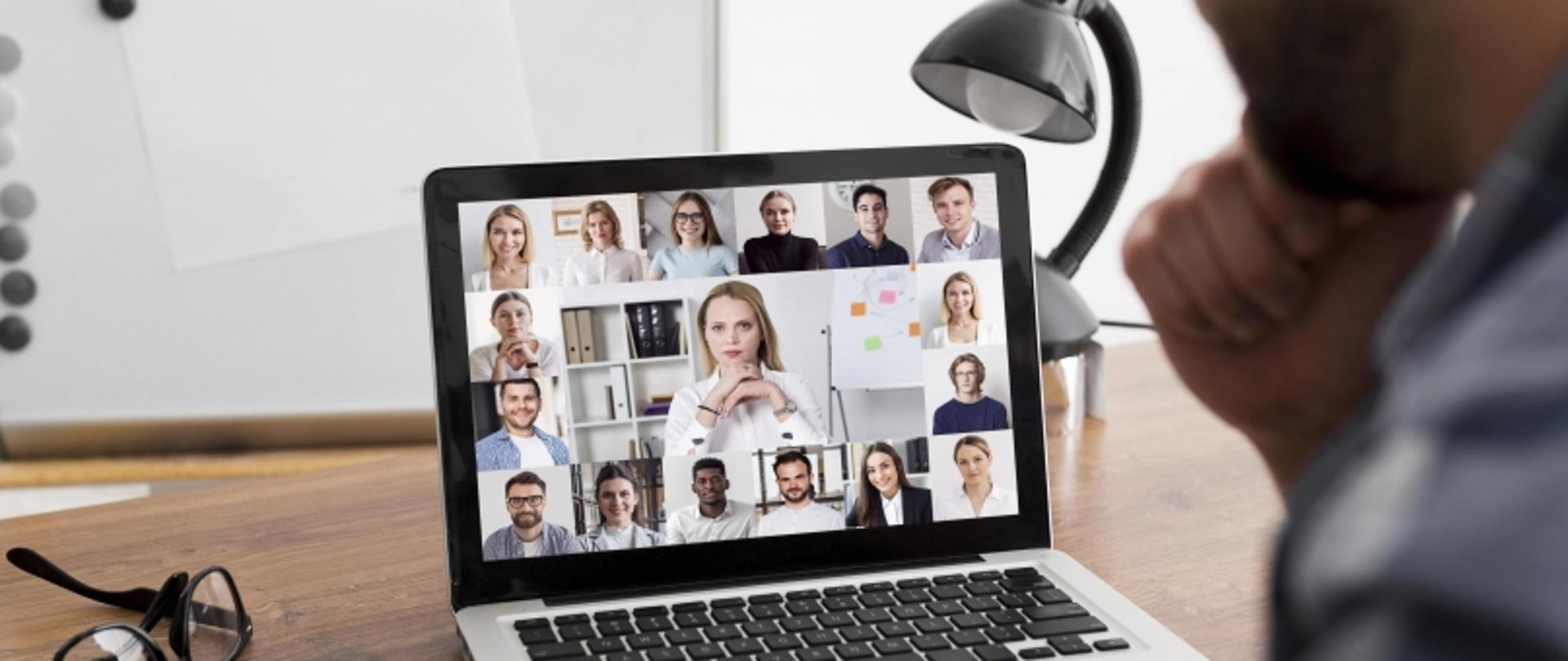 Uczestnik wideokonferencji przy biurku z laptopem, dokumentami i okularami.