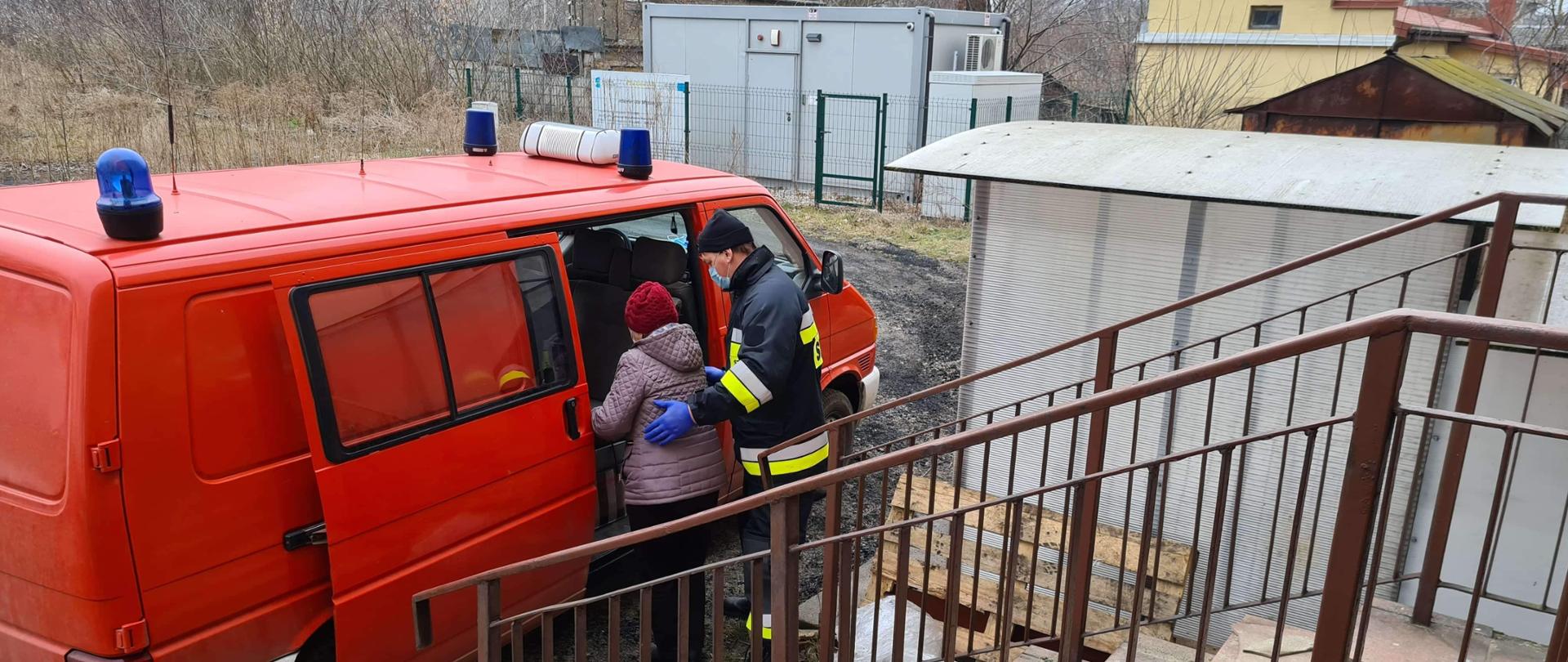 Strażak pomaga starszej kobiecie wejść do samochodu strażackiego.