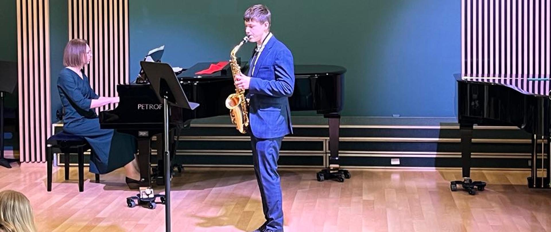 Na zdjęciu widać występ muzyczny w sali koncertowej. Młody mężczyzna w garniturze gra na saksofonie, a kobieta w eleganckiej sukni akompaniuje mu na fortepianie marki Petrof. Na ścianie w tle widnieje logo, które sugeruje, że jest to koncert w Państwowej Szkole Muzycznej (PSM). Sala ma nowoczesny wystrój z drewnianą podłogą i panelami akustycznymi. Publiczność prawdopodobnie obserwuje występ.