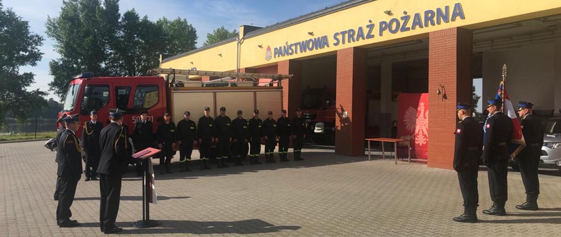 Na zdjęciu widzimy Komendanta Powiatowego, poczet sztandarowy oraz pododdział złożony z funkcjonariuszy Komendy Powiatowej i Jednostki Ratowniczo - Gaśniczej. W tle widzimy pojazd pożarniczy oraz garaże.