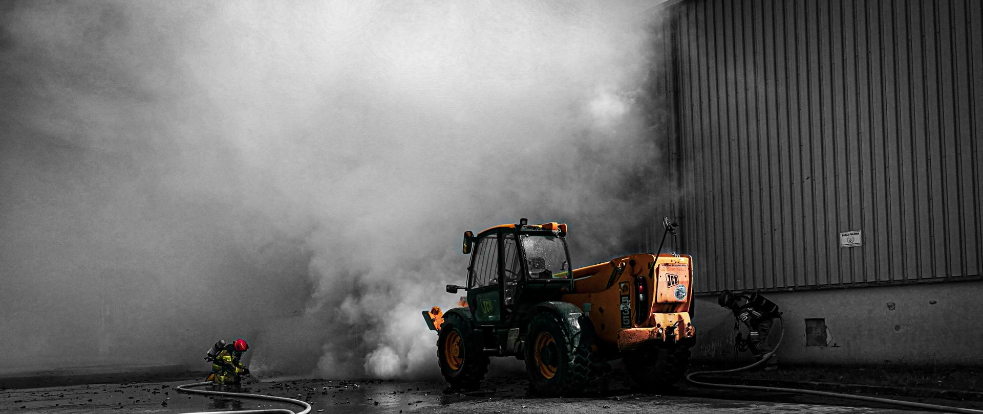 W centrum fotografii znajduje się żółta ładowarka budowlana. Po lewej stronie strażak podaje prąd wody na palącą się ładowarkę. Jest ubrany w ODO. Po prawej stronie przy stojącej obok hali znajduje się strażak, który ma ubrane ODO, a także podaje prąd wody za palącą się ładowarkę. W oddali znajdują się kłęby dymu.
