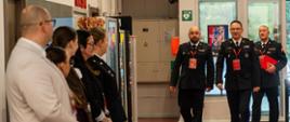 Trzech oficerów Państwowej Straży Pożarnej w mundurach galowych wchodzi do budynku Centralnego Muzeum Pożarnictwa w Mysłowicach. Po lewej stronie stoją osoby oczekujące na ich przybycie.