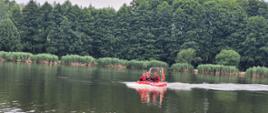 Czterech strażaków ubranych w skafandry płynie łodzią ratowniczą. Za nimi w tle zarosla brzegowe i drzewa liściaste.