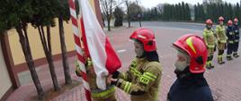 Na pierwszym planie stoi poczet flagowy strażaków w ubraniu specjalnym z zasłoniętymi twarzami i nosem, którzy zakładają Flagę Polski na maszt. Na drugim planie po prawej stronie pozostali strażacy z pododdziału, którzy również są w ubraniu specjalnym z zasłoniętymi twarzami i nosem. Za strażakami znajduje się chodnik i jezdnia. Po lewej stronie widać trzy krzewy. Wydarzenie ma miejsce na placu manewrowym przed Komendą Powiatową. 