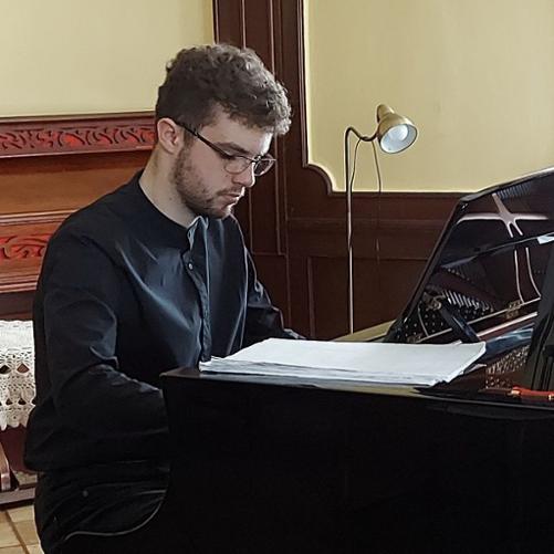 Zdjęcie kolorowe. Młody pianista w ciemnym stroju galowym siedzi przy fortepianie. W tle lampa i fisharmonia.