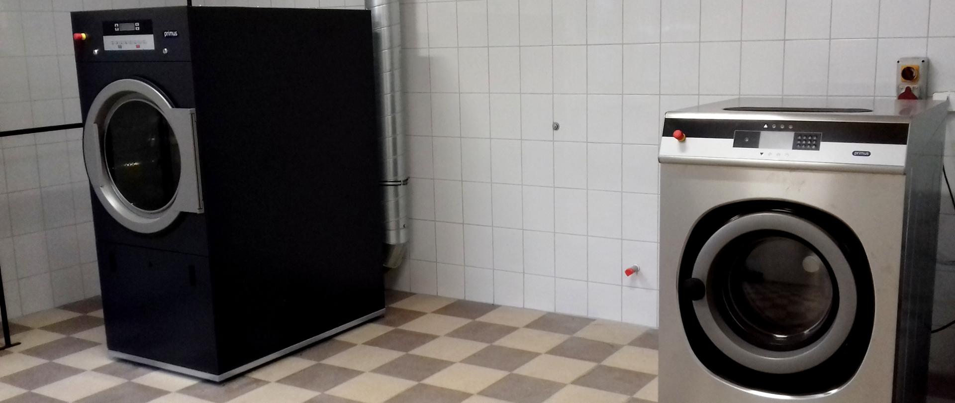Zdjęcie przedstawia pomieszczenie pralni Komendy Miejskiej Państwowej Straży Pożarnej w Krośnie. Na tle ściany wyłożonej białymi płytkami zainstalowana jest srebrna pralnica z dużym otworem na przedniej centralnej ścianie urządzenia na wsad ubrań przeznaczonych do prania. Po lewej stronie pomieszczenia, w pewnej odległości od pralnicy widoczne jest ciemnoszare urządzenie – suszarka do upranych ubrań. W jego przedniej części znajduje się otwór do wkładania odzieży przeznaczonej do suszenia.