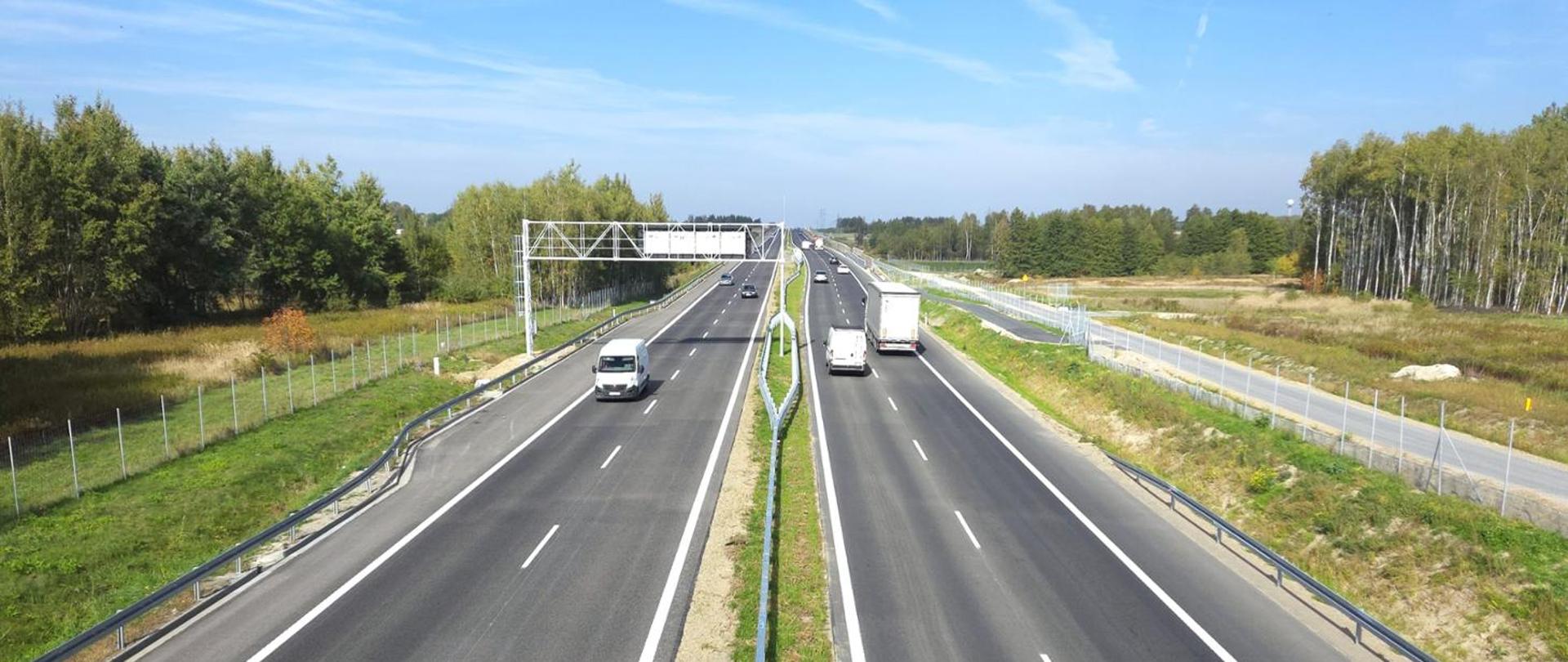 Widok z góry na szeroką, dwujezdniową drogę ekspresową biegnącą przez tereny leśne. Po obu stronach jezdni porusza się kilka samochodów osobowych i ciężarowych. Drogę otaczają zielone pobocza, ogrodzenia oraz las, a nad całością rozciąga się bezchmurne, jasnoniebieskie niebo.