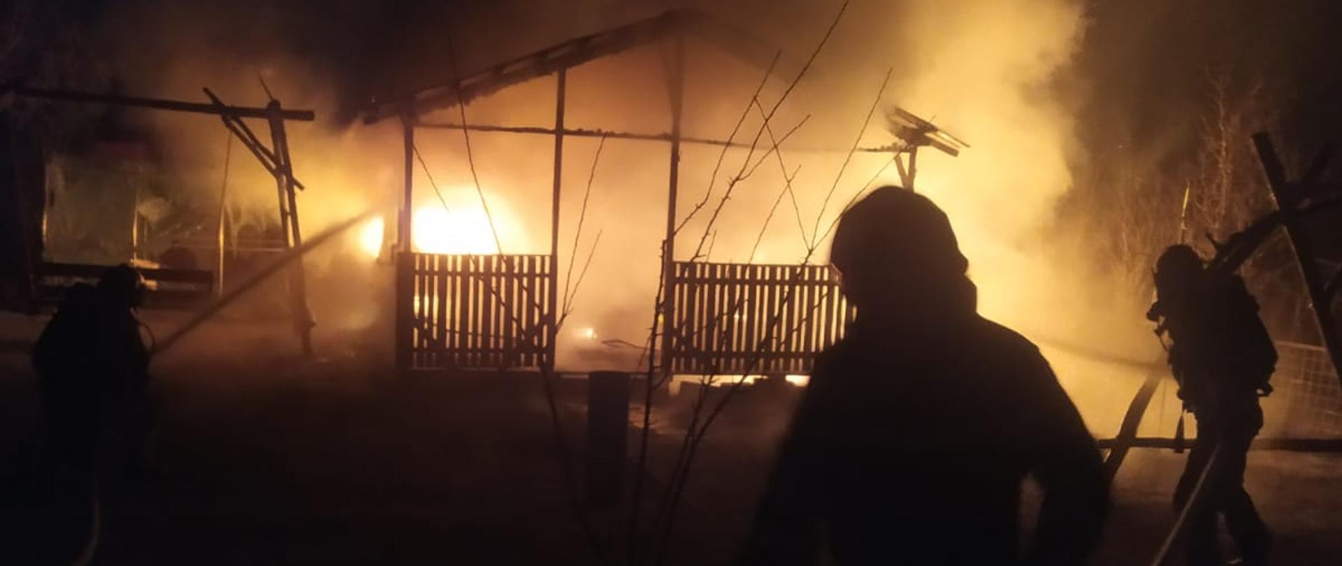 Drewniana altana ogrodowa cała objęta jest ogniem . Pora nocna. Obok altany po lewej stronie widać drewnianą huśtawkę. Strażacy w ubraniu specjalnym w aparatach UODO gaszą palącą się altanę. 