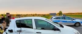 Zdjęcie przedstawia uszkodzony biały samochód marki Renault. Na drugim planie znajdują się osoby postronne oraz policyjny radiowóz.