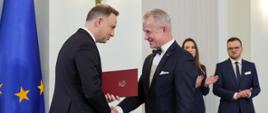 Uśmiechnięty prezydent Duda trzyma czerwoną teczkę i ściska rękę siwemu mężczyźnie w garniturze. Za nimi mężczyzna w garniturze i ubrana na czarno kobieta biją brawo.