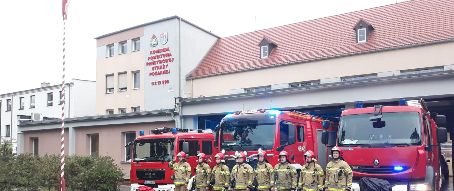 ODDALI HOŁD ZMARŁYM STRAŻAKOM. Na zdjęciu widać strażaków przed budynkiem Komendy, samochody strażackie wyjechały z garażu, mają włączone sygnały świetlne i dźwiękowe, widać maszt z flagą Polski i budynek Komendy Powiatowej PSP w Kościanie