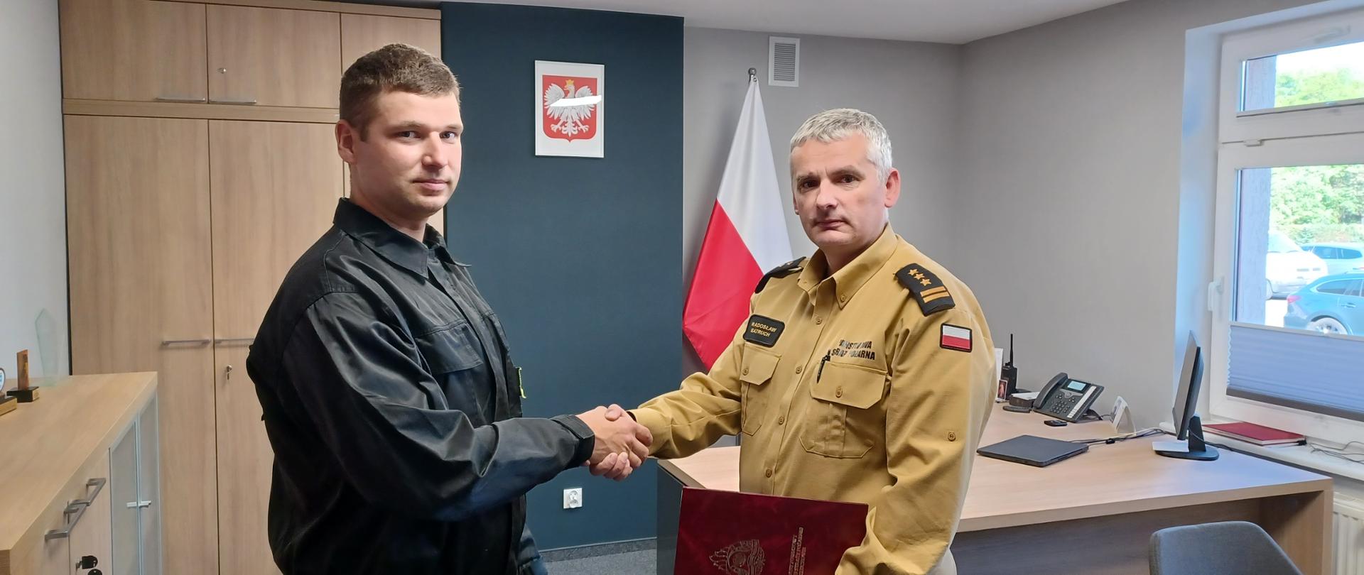 Komendant Powiatowy Państwowej Straży Pożarnej w Człuchowie st. bryg. Radosław Batruch stoi w gabinecie i wręcza akt mianowania na stanowisko dowódcy zmiany st. asp. Dariuszowi Buczko.