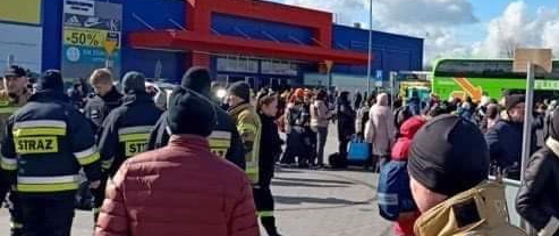 Zdjęcie przedstawia plac przy Tesco. Strażacy koordynują przemieszczanie się uchodźców do autobusów przewożących ich do punktów docelowych w Polsce i za granicą.