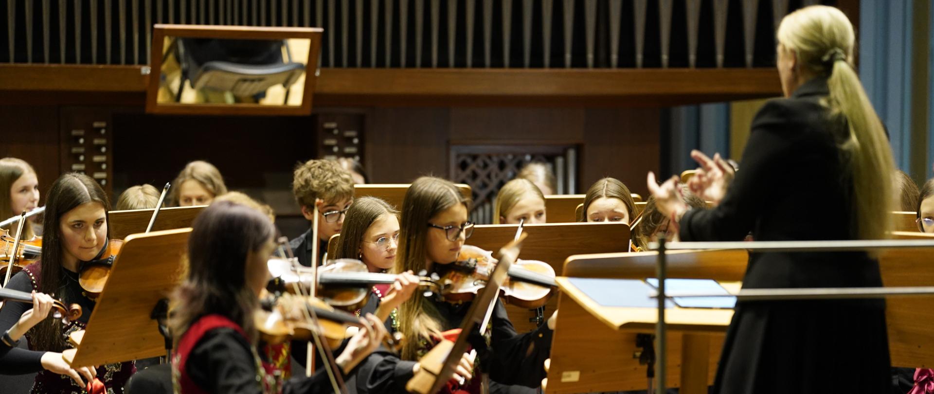 Zdjęcie przedstawia młodzieżową orkiestrę smyczkową podczas występu lub próby w sali koncertowej. Oto główne elementy widoczne na fotografii: Muzycy: Na pierwszym planie widać młode instrumentalistki grające na skrzypcach. Są ubrane w eleganckie, ciemne stroje z charakterystycznymi, tradycyjnymi elementami dekoracyjnymi (haftowane kamizelki z motywami kwiatowymi i czerwone wstążki), co sugeruje inspirację folklorem. Dyrygentka: Po prawej stronie, tyłem do obiektywu, stoi kobieta dyrygująca zespołem. Ma długie jasne włosy spięte w kucyk i jest ubrana w czarną suknię lub płaszcz. Otoczenie: W tle widoczne są drewniane pulpity na nuty oraz monumentalne organy piszczałkowe, które stanowią tło sceny. Nad orkiestrą zawieszone jest lustro, prawdopodobnie ułatwiające muzykom kontakt wzrokowy z dyrygentem lub organistą. Atmosfera zdjęcia jest pełna skupienia i profesjonalizmu, typowa dla uroczystego wydarzenia muzycznego.