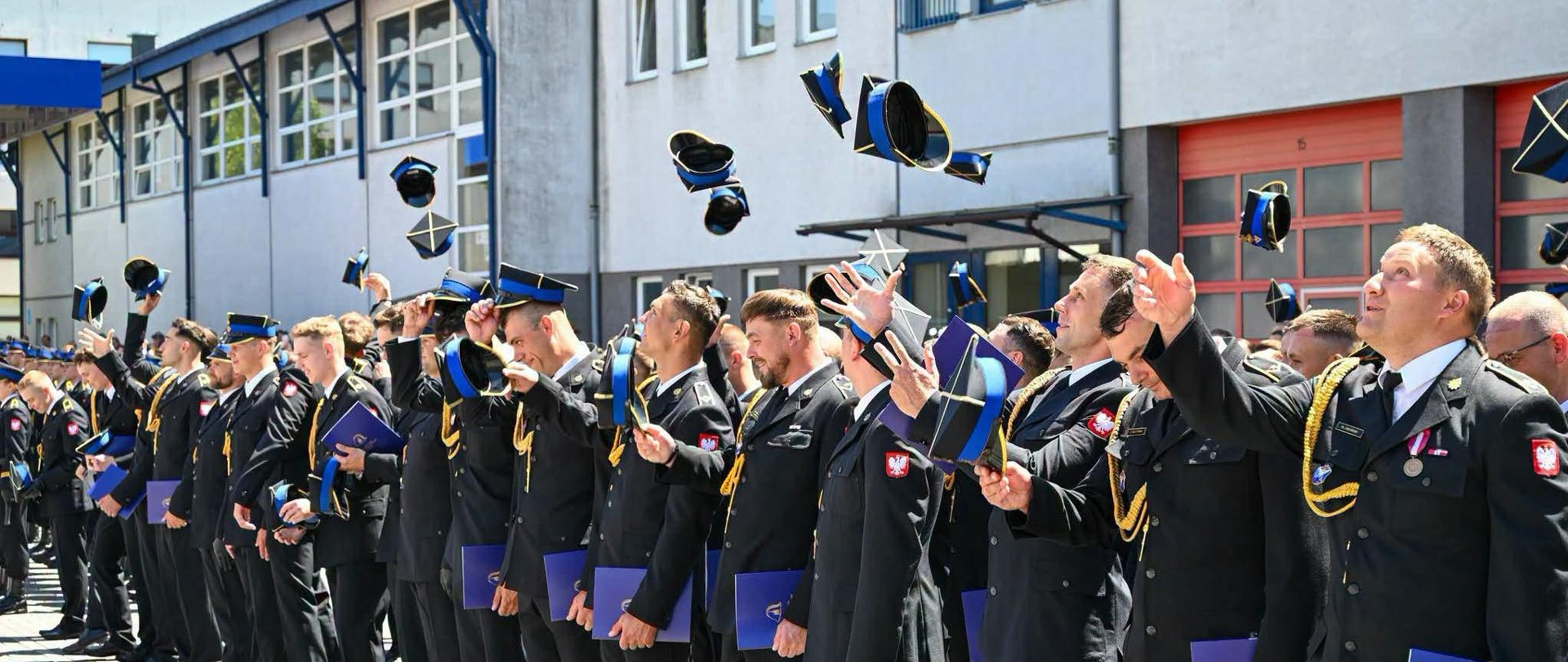 Grupa strażaków Państwowej Straży Pożarnej w galowych mundurach stoi w szeregu na placu przed budynkiem komendy. Wszyscy z uśmiechem i radością podrzucają w górę swoje czapki rogatywki podczas uroczystego apelu. W rękach trzymają niebieskie foldery lub dyplomy. W prawym dolnym rogu widnieje podpis: „st. str. kdt Piotr Krzak | Państwowa Straż Pożarna”"
