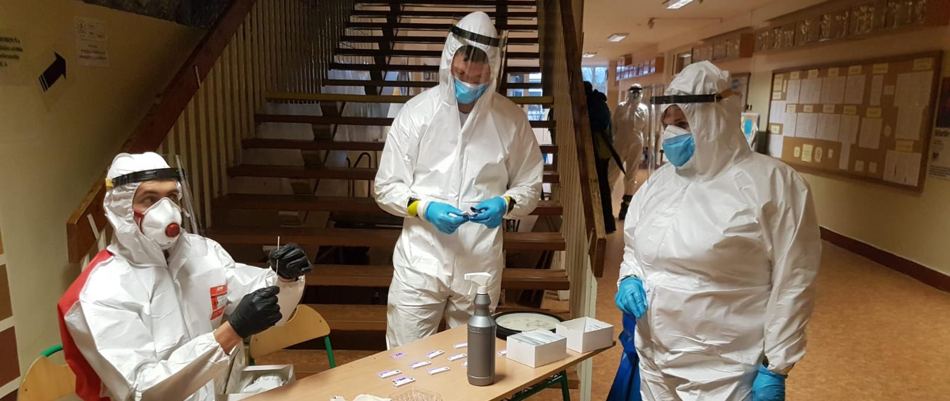 zdjęcie przedstawia trzech strażaków w środku budynku szkoły, którzy są ubrani w białe ubrania ochrony indywidualnej , maseczki, przyłbice i rękawiczki. Strażacy gotowi są do pobrania wymazów na COVID-19. W oddali widać strażaka ubranego w środki ochrony indywidualnej, który czeka na przychodzących ludzi.