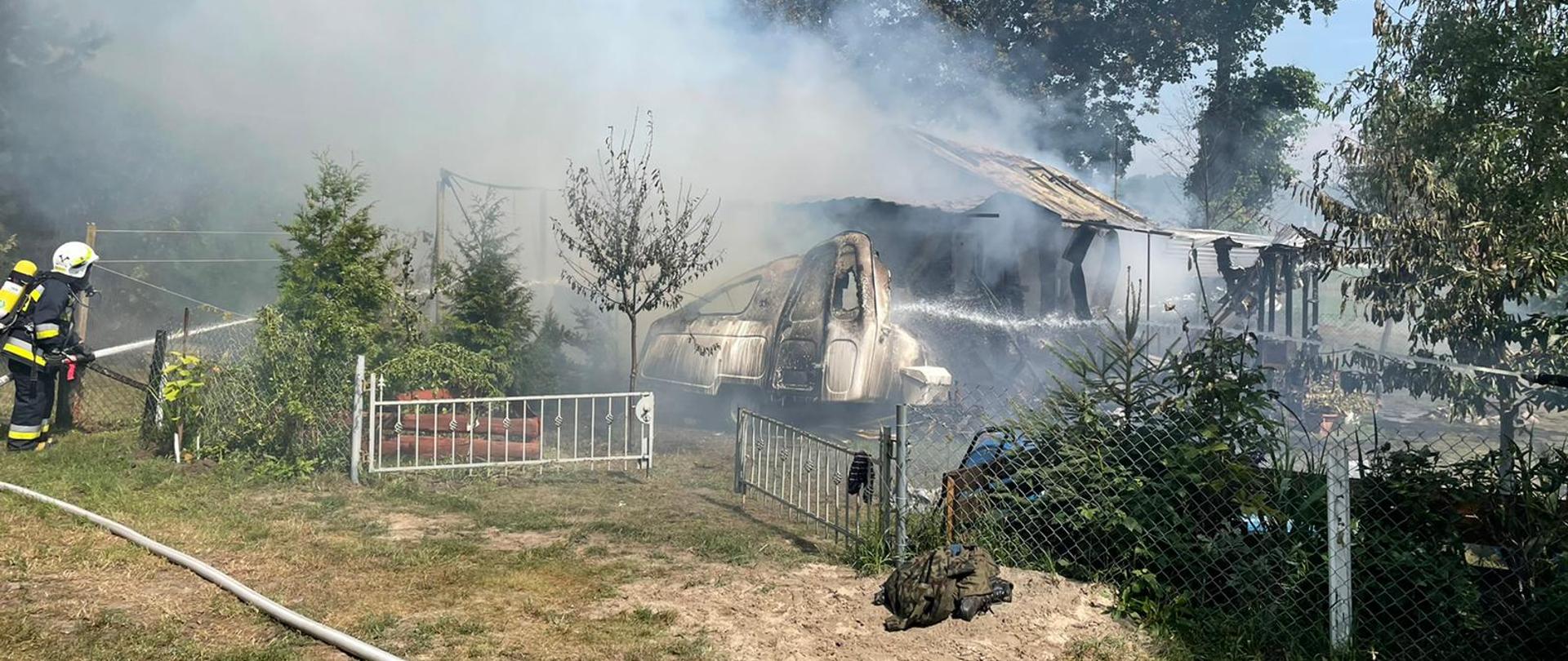 Na zdjęciu pożar altany, krzewy oraz drzewa z lewej strony strażak podający prąd wody na palącą altanę
