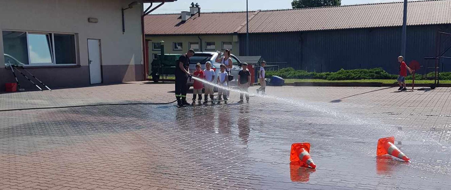 Dzieci wraz ze strażakiem lejące wodę z prądownicy do pachołków. po lewej stronie garaż JRG. W tle budynki. 