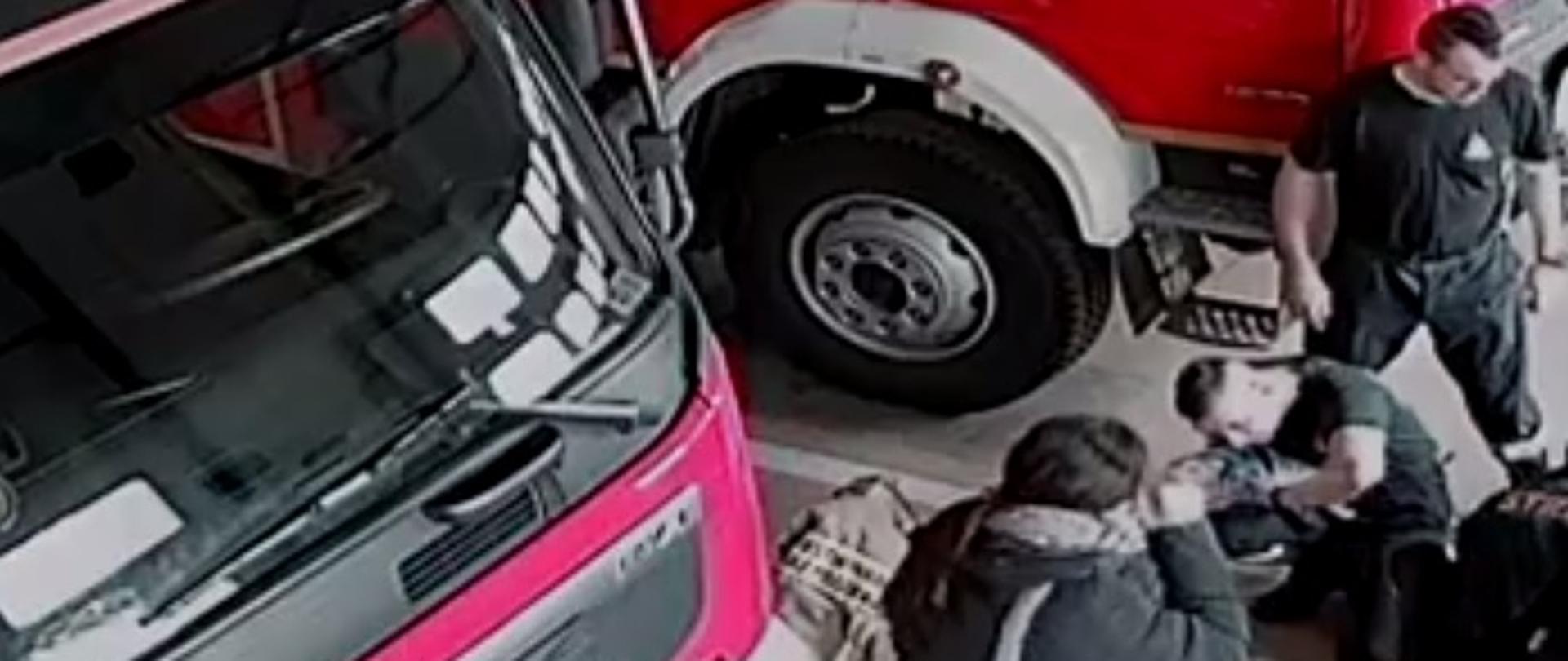 Zdjęcie obrazuje strażaków i inną osobę oraz pojazdy pożarnicze