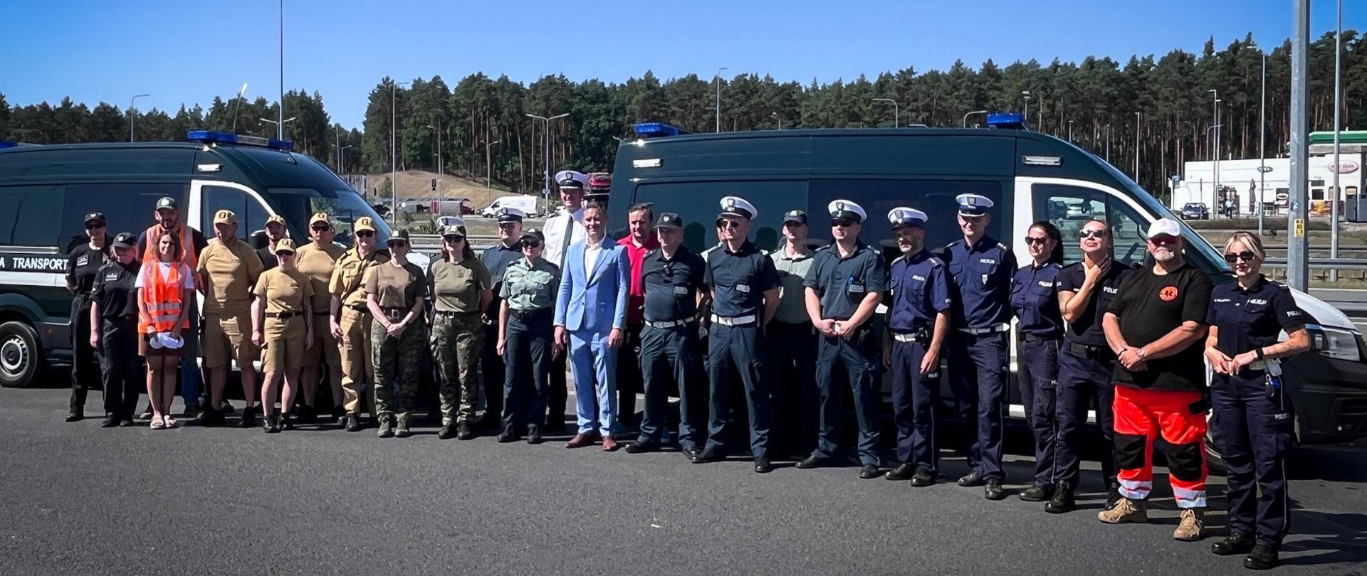 Pamiątkowe zdjęcie grupowe uczestników ćwiczeń na placu lub parkingu. W centrum stoi mężczyzna w jasnoniebieskim garniturze, a wokół niego zgromadzili się funkcjonariusze w różnych mundurach: policjanci drogówki w białych czapkach, policjanci w granatowych garnizonowych mundurach, żołnierze w kamuflażu, przedstawiciele Inspekcji Transportu Drogowego (ITD) oraz ratownicy medyczni. W tle stoją dwa policyjne busy transportowe, a za nimi rozciąga się ściana lasu i wysokie latarnie uliczne w pełnym słońcu.