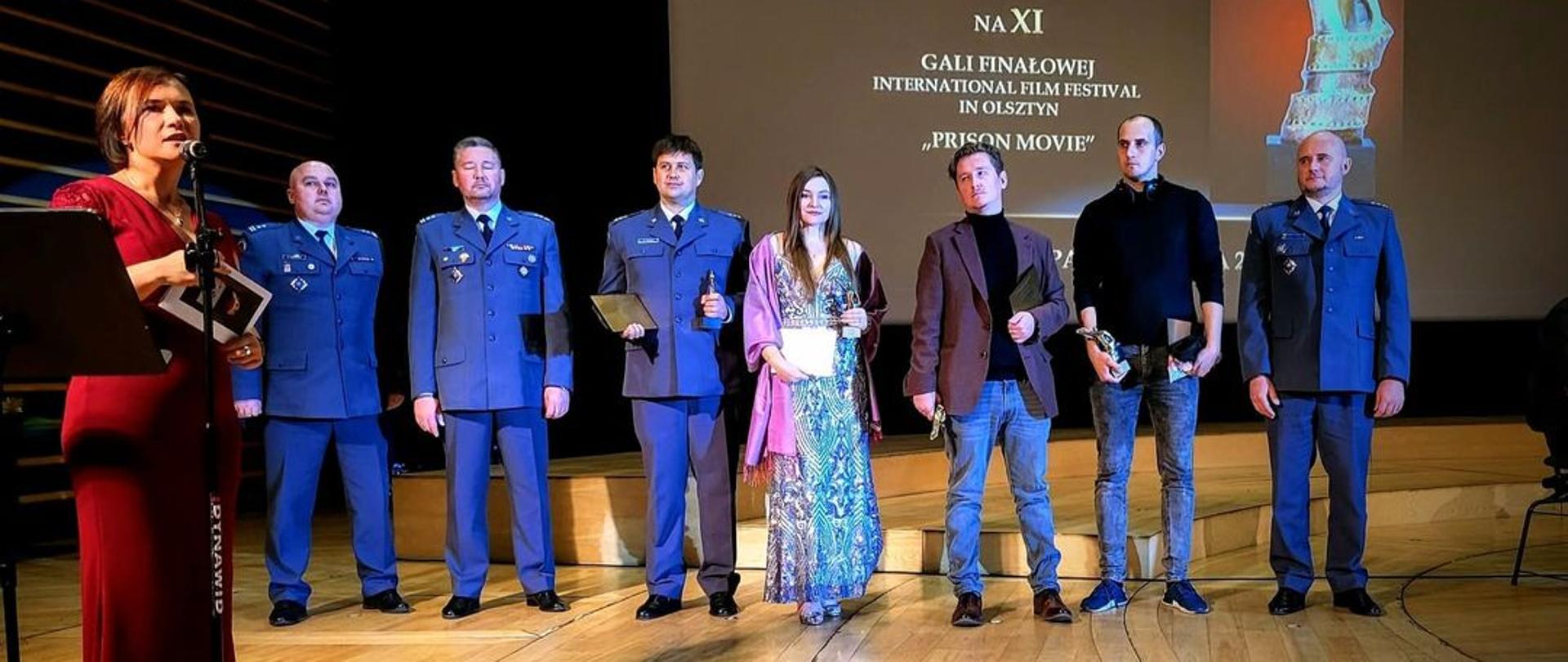 Scena filharmonii w Olsztynie. Prezenterka ubrana w czerwoną długą sukienkę. Siedmiu laureatów Prison Movie Festival ustawionych w rzędzie trzymając statuetki. Czterech z nich w mundurach Służby Więziennej. Z tyłu na ścianie na projektorze wyświetlony slajd prezentacji o gali finałowej.