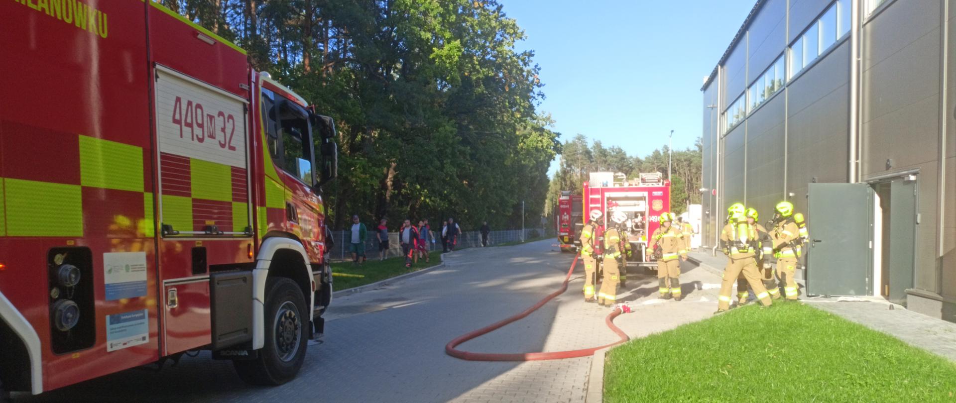 Zdjęcie przedstawia ćwiczenia straży pożarnej na zewnątrz. Na pierwszym planie, z lewej strony, widoczny jest czerwony wóz strażacki z Milanówka (oznaczenie 449M32). Po prawej stronie, na trawniku przed szarą halą przemysłową, grupa pięciu strażaków w pełnym umundurowaniu (żółto-brązowe kombinezony, zielone kaski) obsługuje czerwony wąż strażacki, który leży rozciągnięty na brukowanym chodniku. W tle widać drugi wóz strażacki i grupę obserwatorów. Tło stanowi gęsty las pod błękitnym niebem.