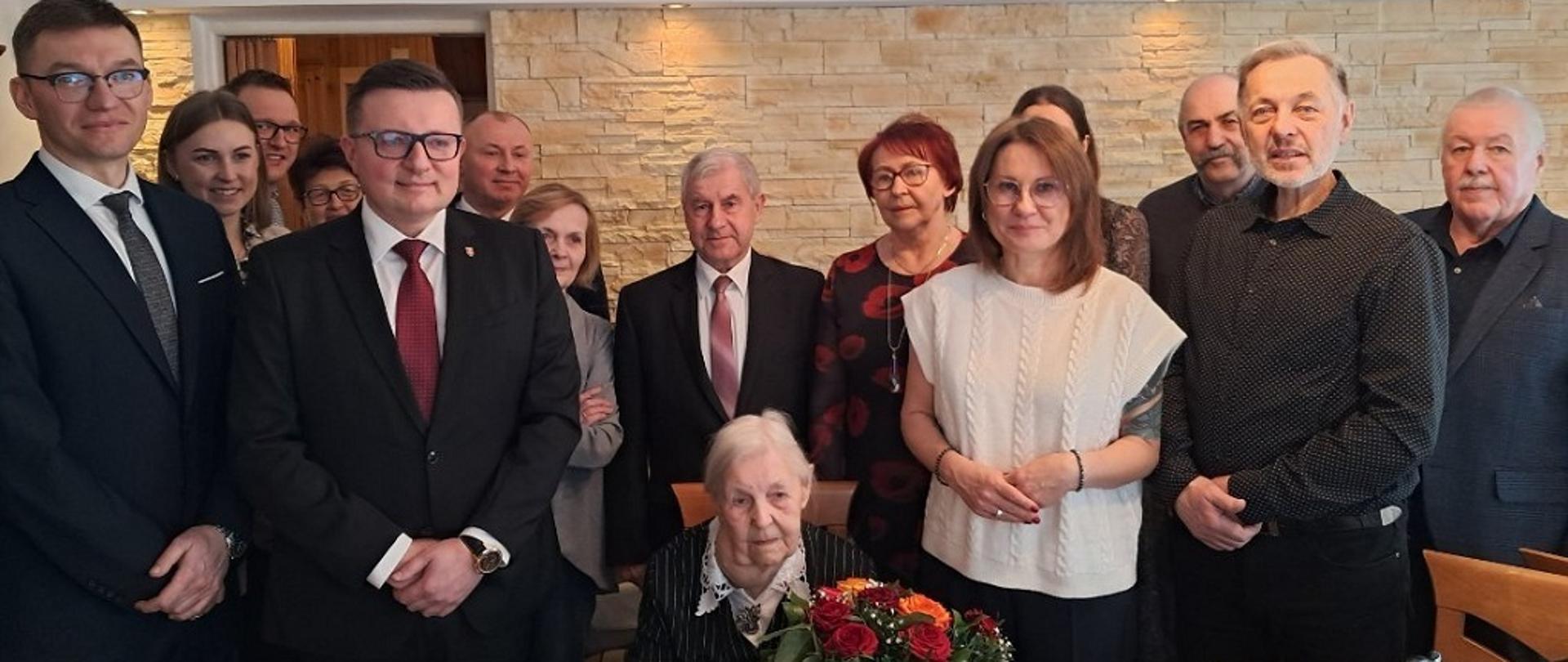 Na zdjęciu jest 8 mężczyzn i 7 kobiet. Po środku siedzi na krześle kobieta i trzyma bukiet kwiatów
