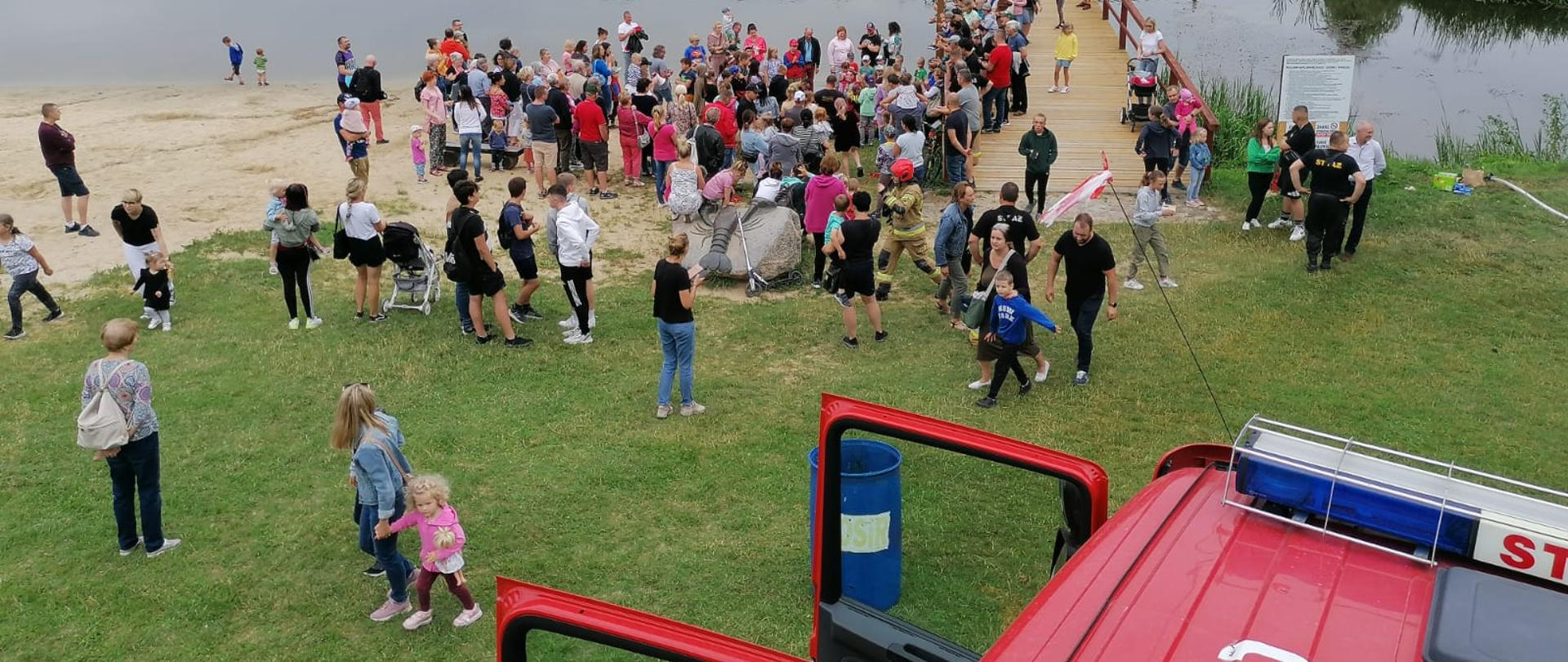 Dzieci z rodzicami zgromadzeni przy pomoście nad jeziorkiem, w Parku Ekologicznym, w Pasłęku. Z prawej strony widoczny dach pojazdu pożarniczego.