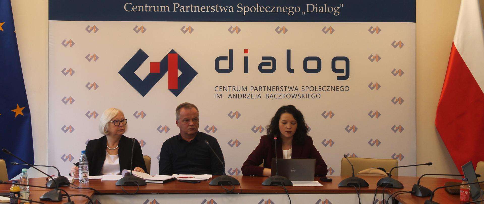 Na zdjęciu widnieje stół prezydialny za którym siedzi wiceminister Rodziny, Pracy i Polityki społecznej – Katarzyna Nowakowska, która prowadzi spotkanie. W tle znajduje się baner z napisem Centrum Partnerstwa Społecznego Dialog. Od lewej strony zasiadają współprzewodnicząca Zespołu ze strony pracodawców – Teresa Hernik oraz Zbigniew Spendel – współprzewodniczący strony związkowej. 