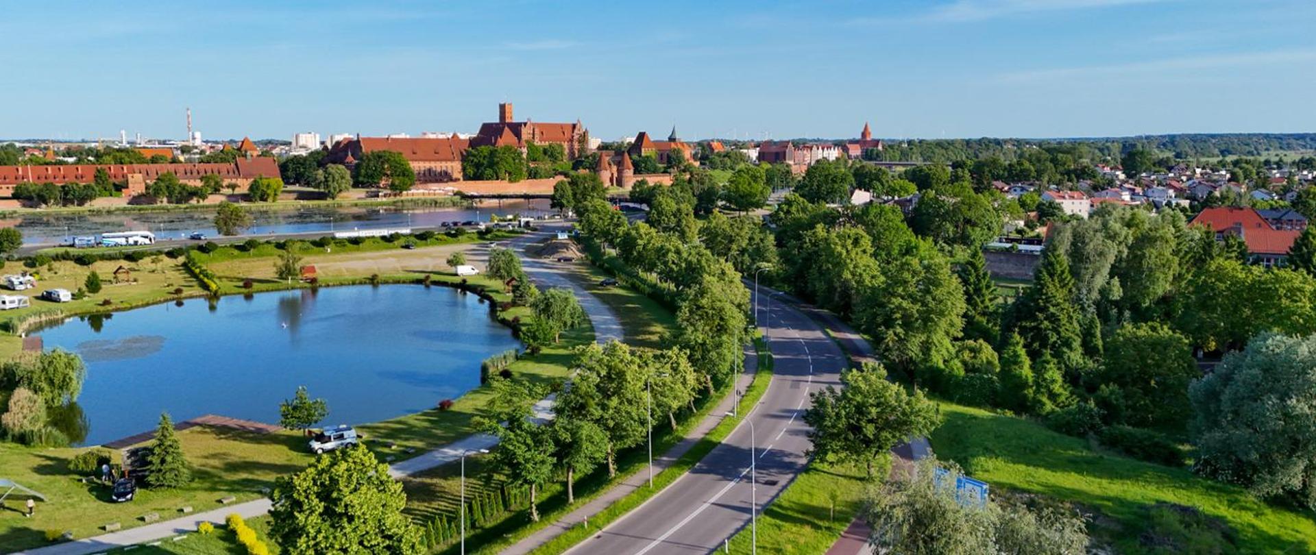 Widok z lotu ptaka na drogę biegnącą wśród zieleni i stawów w kierunku miasta. W tle widoczna zabytkowa zabudowa z charakterystycznym zespołem ceglanych budynków nad rzeką, otoczona drzewami i terenami zielonymi.