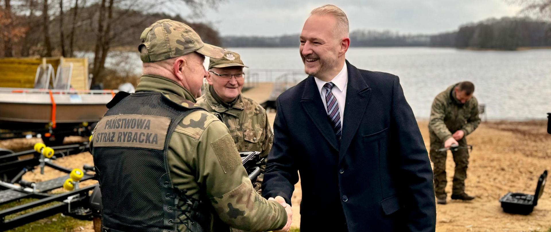 Uroczyste przekazanie sprzętu Państwowej Straży Rybackiej w Olsztynie w dniu 5 grudnia 2024 r. fot. nr 5. wojewoda podaje rękę i przekazuje sprzęt. W tle jezioro Skanda. 
