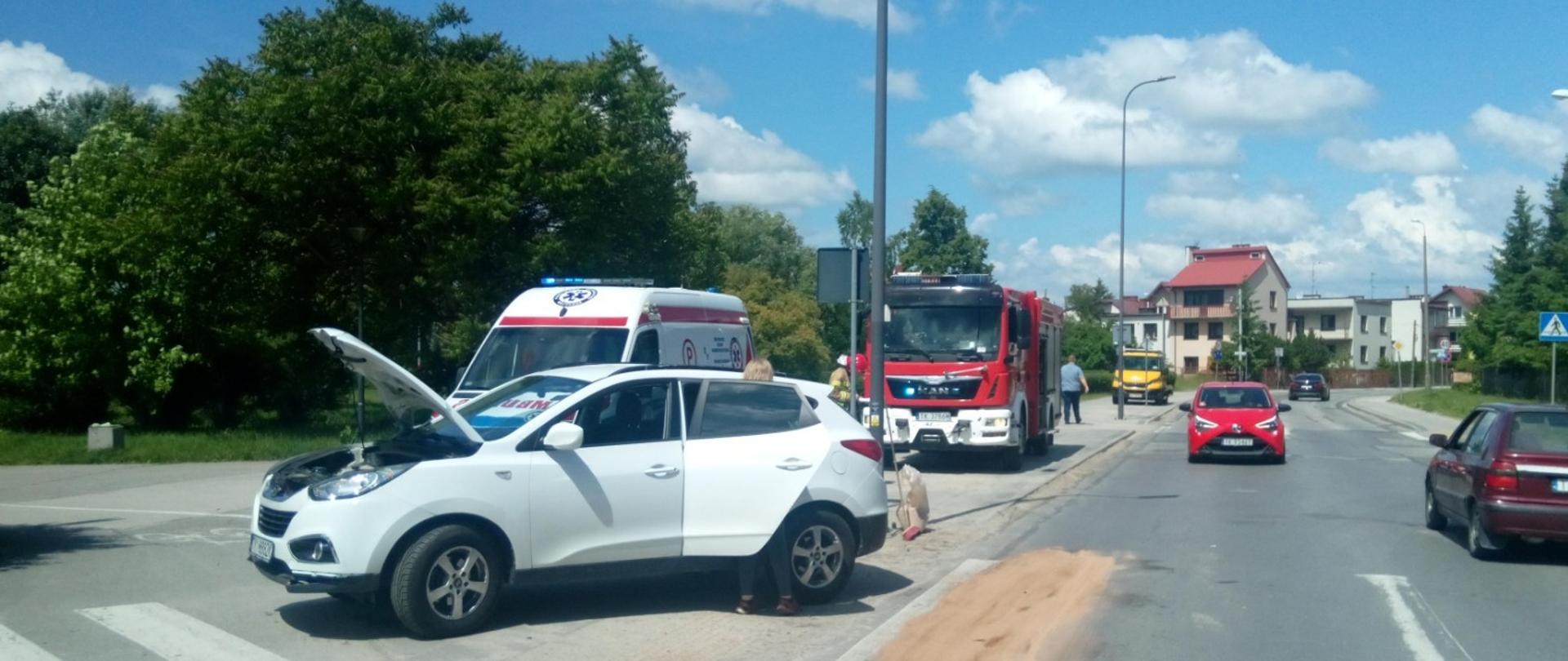 Zdjęcie przedstawia Aleję Legionów w Kielcach. Na pierwszym planie samochód osobowy biały Hyundai, który brał udział w kolizji drogowej. Za nim widać stojący ambulans i wóz strażacki. Na jezdni duża plama rozsypanego sorbentu.