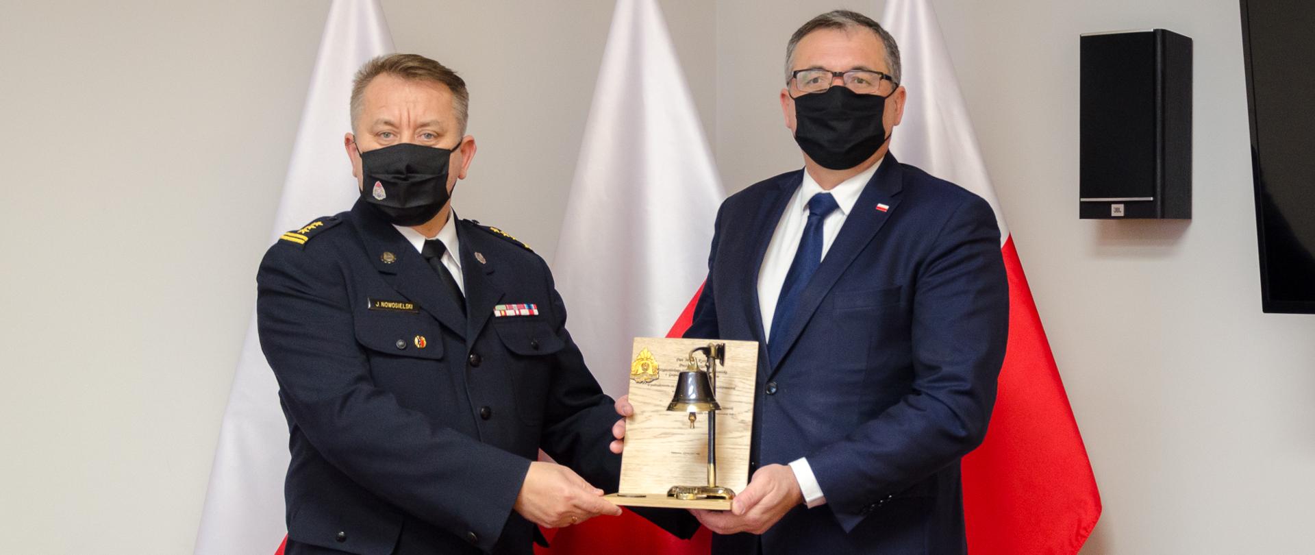 Zdjęcie przedstawia dwie osoby, strażaka i osobę w ubraniu cywilnym które stoją na tle trzech flag Polski. W dłoniach trzymają okolicznościową statuetkę w kształcie toporka z dzwonkiem.

