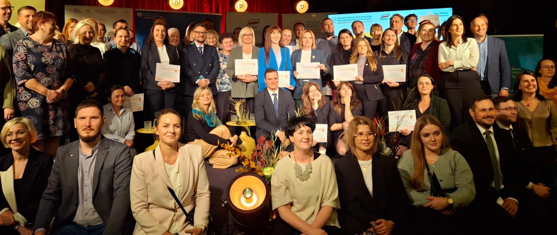 Szeroki kadr. Zdjęcie grupowe. Uczestnicy wydarzenia pozują wspólnie, część osób trzyma w dłoniach certyfikaty Twórców Społeczeństwa Obywatelskiego