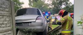 Na betonowym ogrodzeniu zawieszony jest samochód osobowy wokół którego prowadzą działania strażacy.