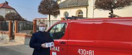 Na tle czerwonego strażackiego busa na pierwszym planie stoi ksiądz w sutannie trzymający pudełko z maseczkami. Za samochodem żółty kościół.