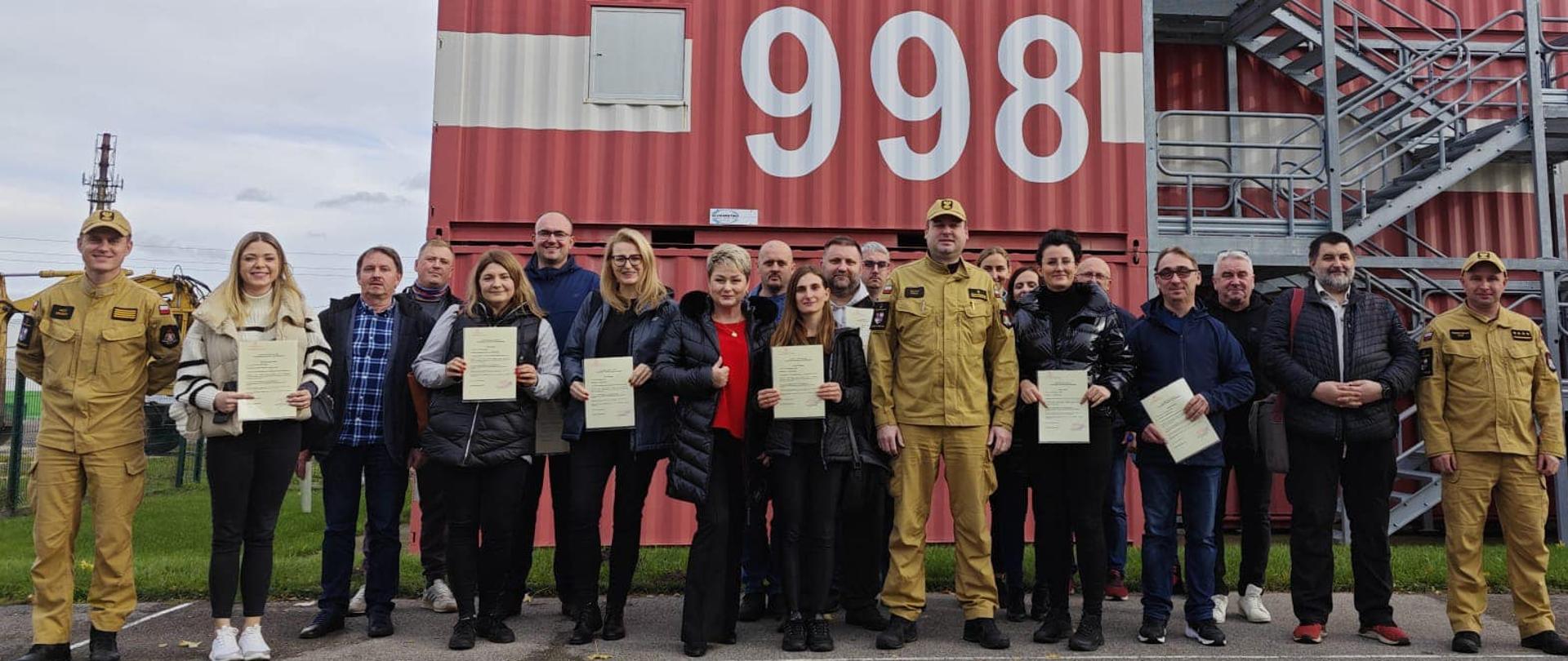 Na zdjęciu widzimy grupę szkoleniową wraz z wykładowcami na poligonie przed kontenerami treningowymi PSP. Kursanci prezentują w rękach świadectwa ukończenia kursu.