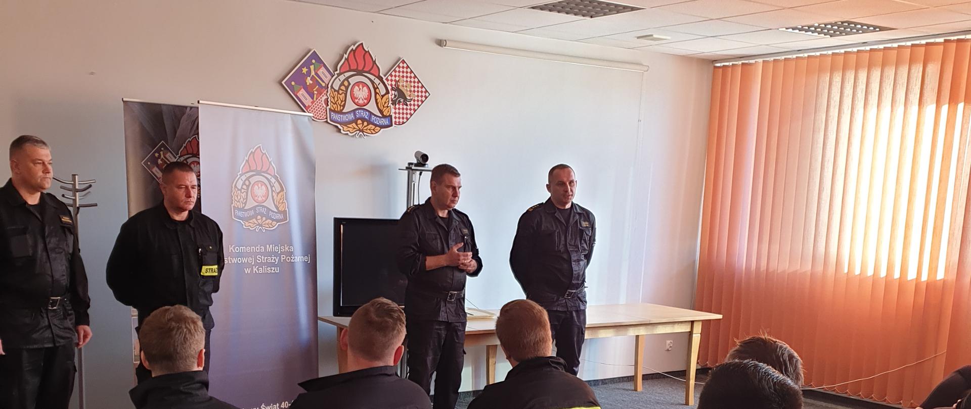 Zdjęcie zrobione w sali. Druhowie siedzą na krzesłach. Osoby te są umundurowane. Przed nimi stoją funkcjonariusze PSP, umundurowani. Rolety w oknach. Dywan na podłodze, ściany żółte. Na ścianie widoczne logo PSP.