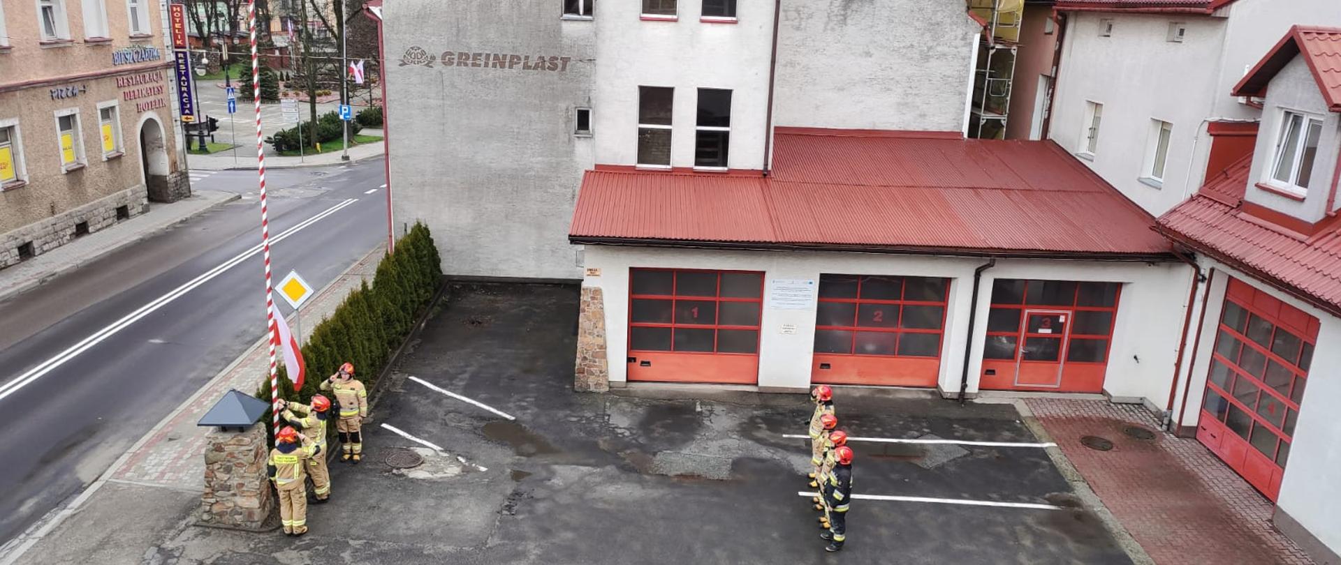 Na zdjęciu widać zmianę służbową ubraną w ubranie specjalne na placu komendy. Czworo strażaków stoi na wprost masztu przy którym jest kolejnych 3 strażaków którzy uroczyście podnoszą flagę Rzeczypospolitej Polskiej na maszt. Na zdjęciu widać jeszcze drogę główną oraz garaże komendy PSP w Ustrzykach Dolnych